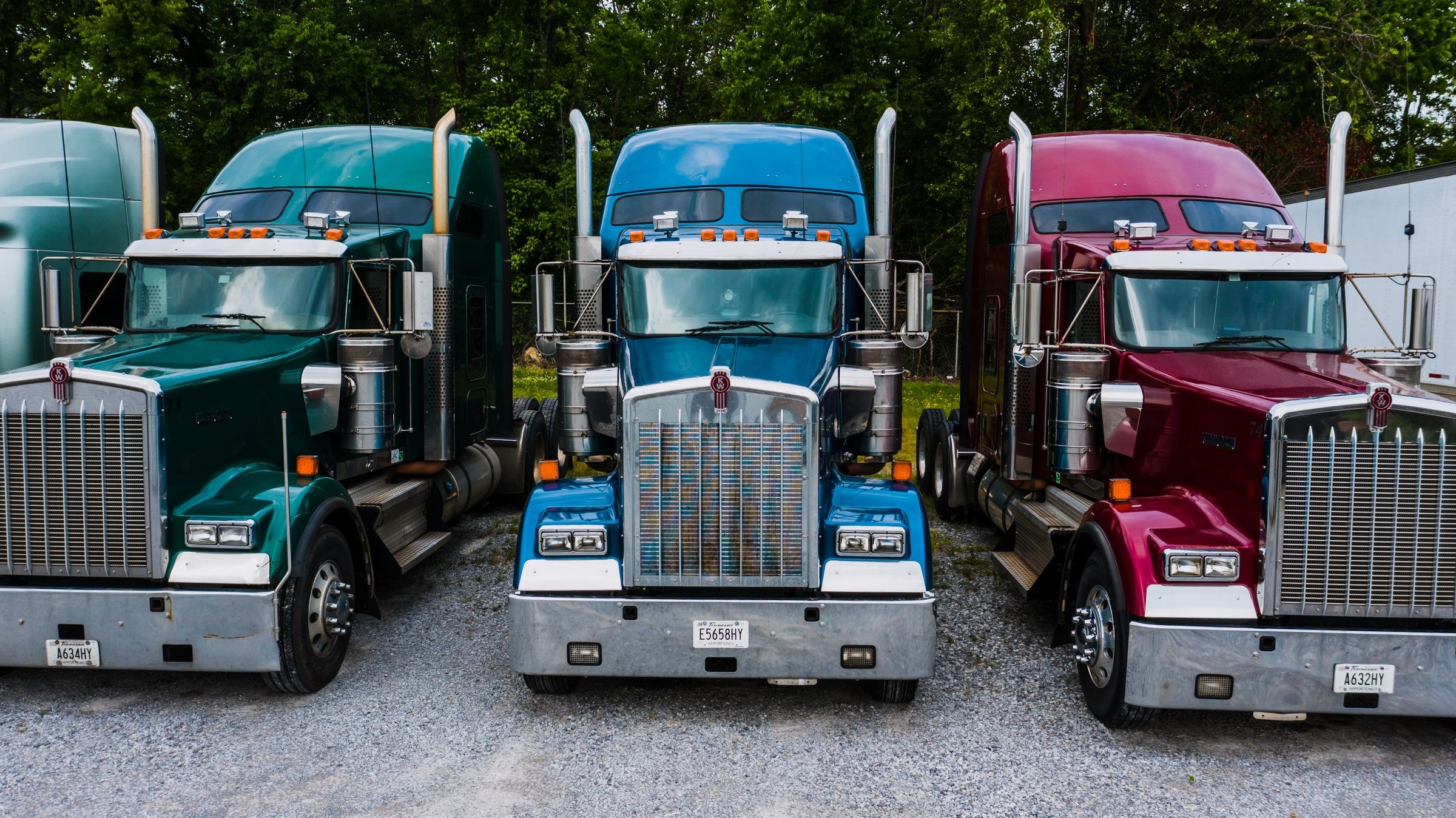 American semi-truck cabs parked at a tree-lined gravel depot.