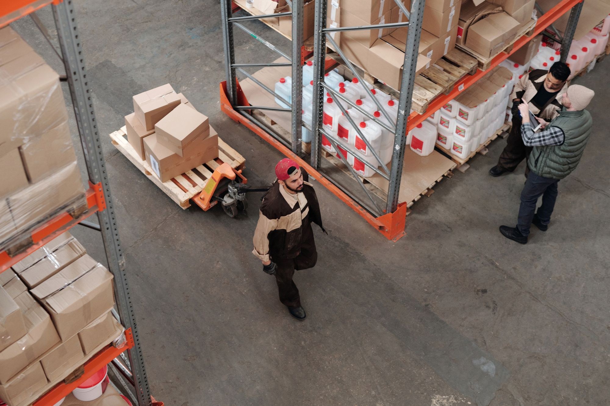 A warehouse worker pulls a pallet jack loaded with cartons while colleagues check inventory.